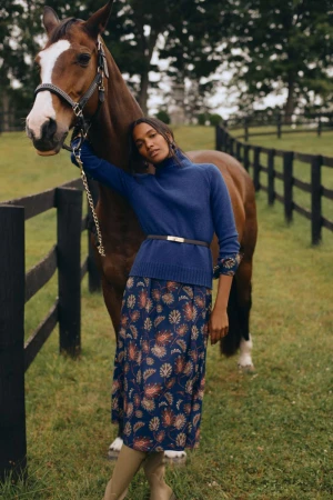 Woman in blue turtleneck and patterned skirt poses with a brown horse near a black wooden fence in a grassy, outdoor environment.