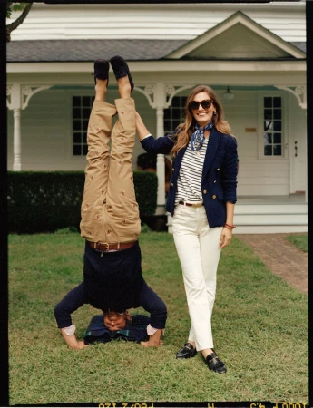 Two people in preppy attire pose on a lawn with a white house in the background. One person is doing a handstand.