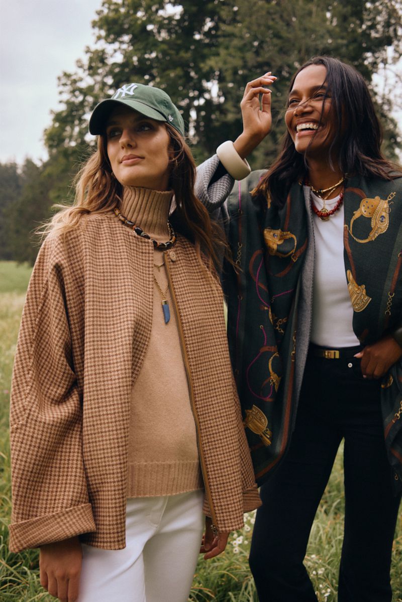 Two women in an open field. One with a baseball cap and jacket, the other with a printed shawl, laughing in natural light.