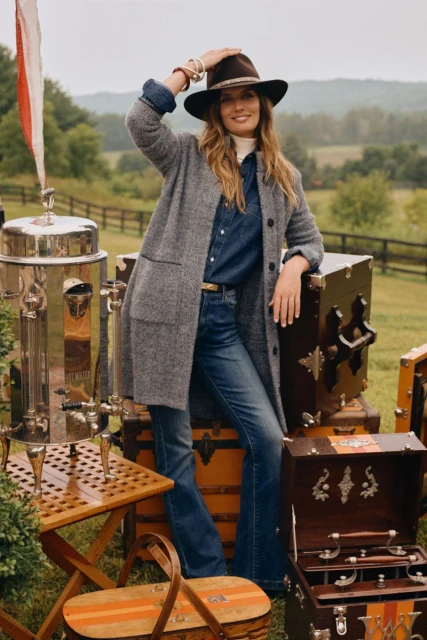 Adult woman in a gray tweed coat poses outside near vintage trunks and green field. She wears a hat, denim shirt, and jeans.