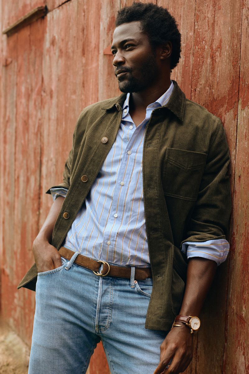 Portrait of a Black man in casual fashion against rustic red wall. He wears a button-down shirt, jacket, jeans, and a brown leather belt.