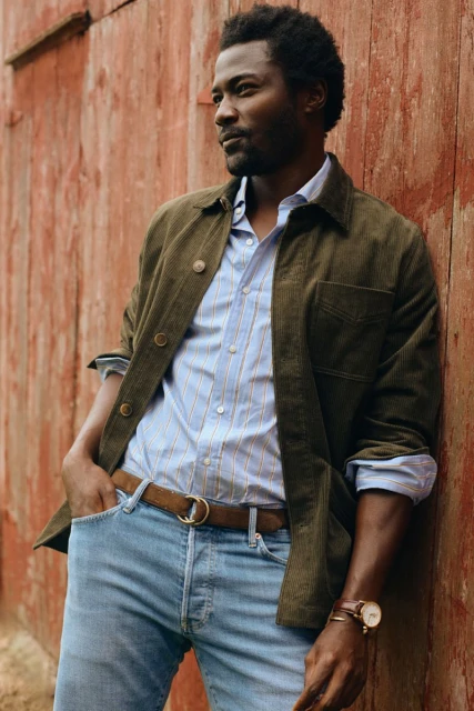 Portrait of a Black man in casual fashion against rustic red wall. He wears a button-down shirt, jacket, jeans, and a brown leather belt.