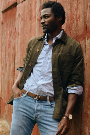 Portrait of a Black man in casual fashion against rustic red wall. He wears a button-down shirt, jacket, jeans, and a brown leather belt.