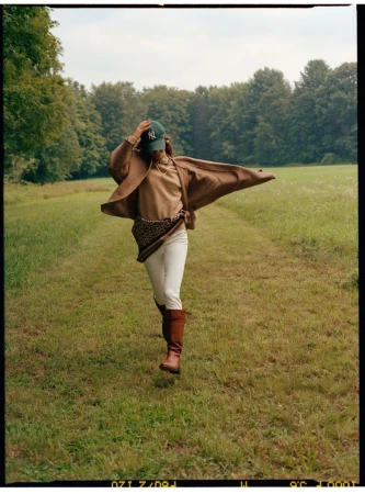 Woman walking on a grassy path wearing a brown cardigan, white pants, tall boots, and a green baseball cap.