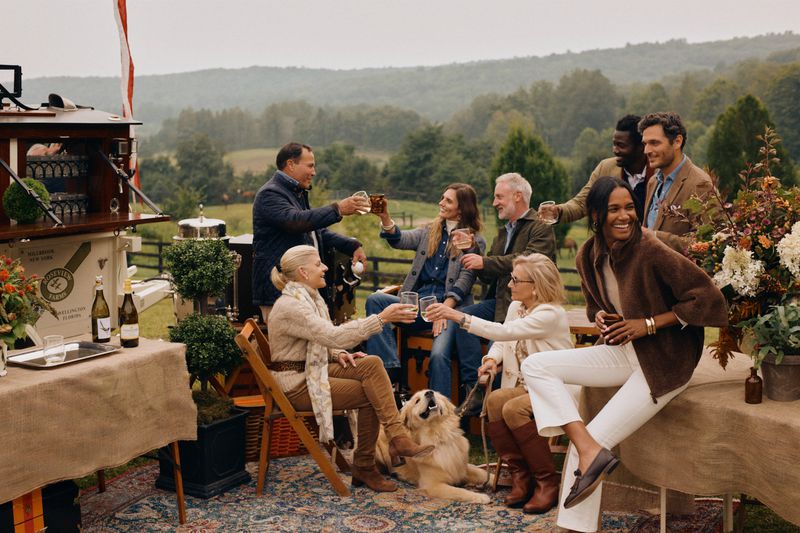 A group of adults gather outdoors on a large lawn for a social gathering while toasting glasses; a dog rests nearby.