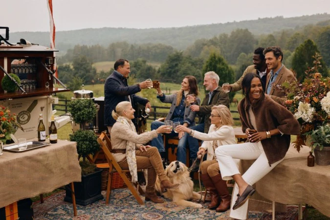 A group of adults gather outdoors on a large lawn for a social gathering while toasting glasses; a dog rests nearby.