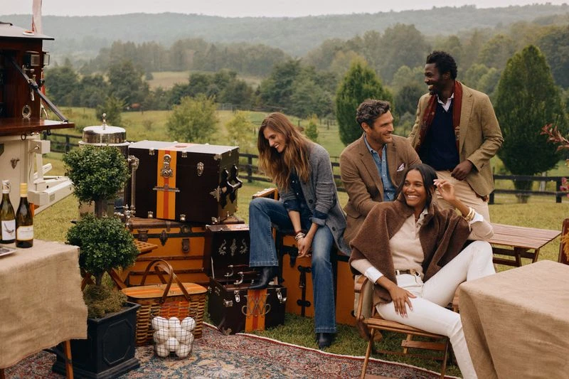 A group of four people are enjoying an outdoor gathering on a grassy field, sitting and standing around vintage trunks and picnic setups.