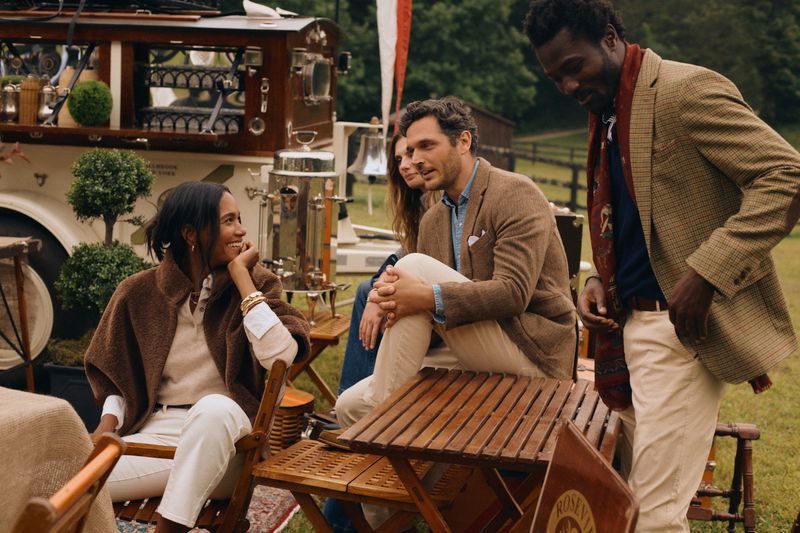 Four adults in tailored jackets and cream pants sit and stand outside an outdoor cafe with a vintage food truck and green lawn.
