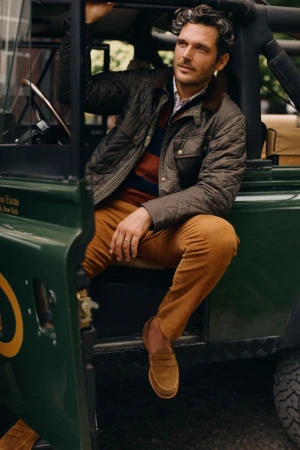 Man seated in a green off-road vehicle wearing a quilted olive jacket, striped sweater, and brown trousers.