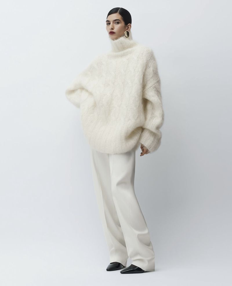 Fashion shoot features a woman in an oversized cream sweater and white pants posing in a light studio with a minimalist aesthetic.
