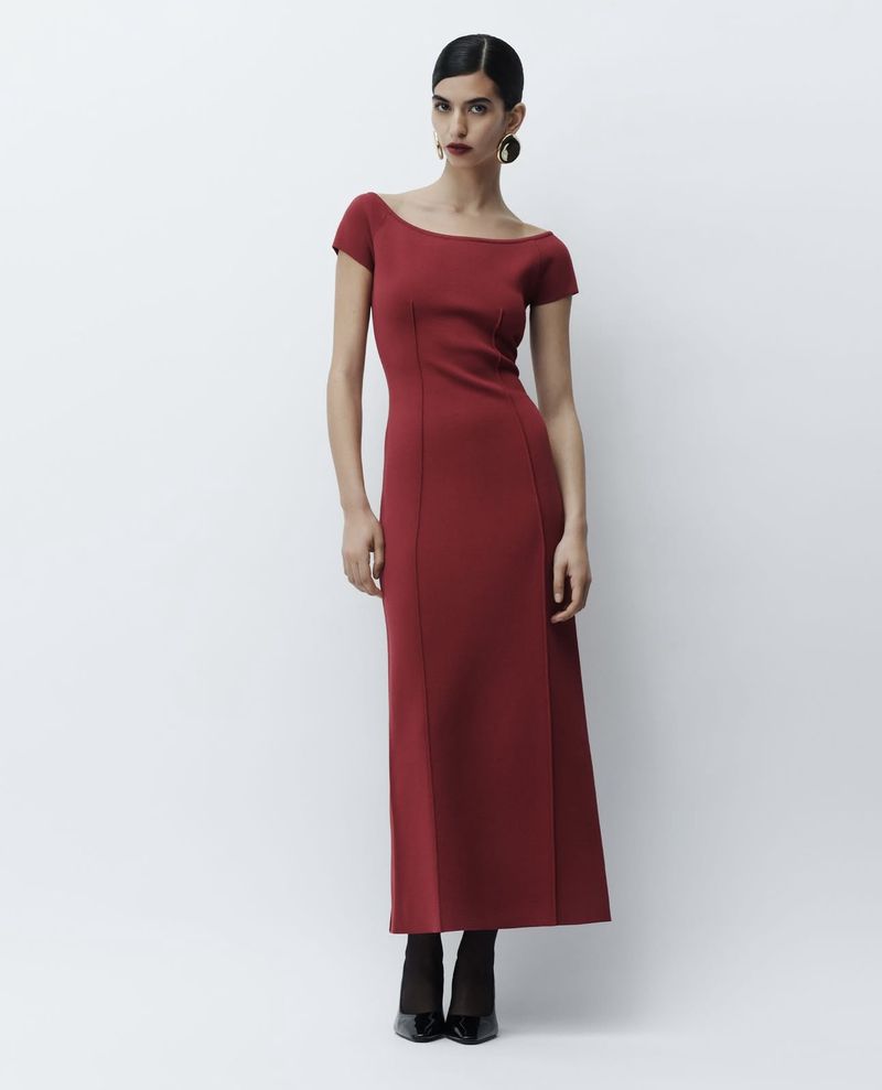 A woman poses in a long, fitted red dress with short sleeves against a white background, showcasing a blend of fashion and simplicity.