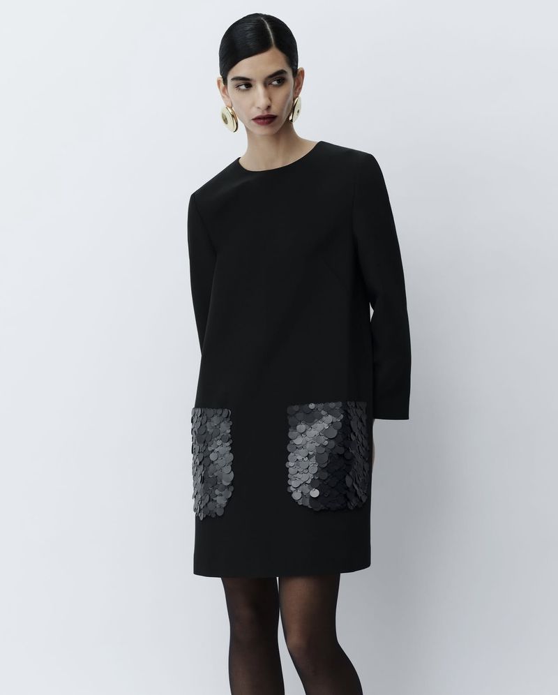A brunette woman is posing in a knee-length black dress with shiny black paillette pocket details on a light grey backdrop.