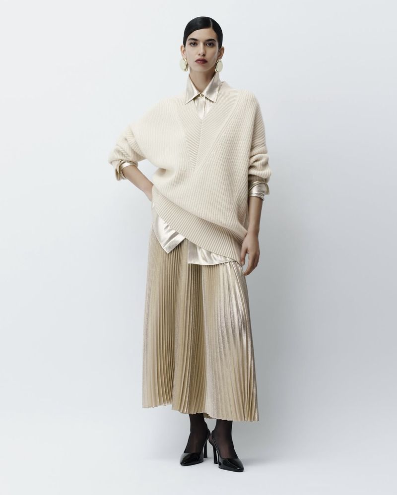 Indoor studio shot of a female fashion model wearing a v-neck sweater with a shiny gold pleated skirt and black heels, posing gracefully.
