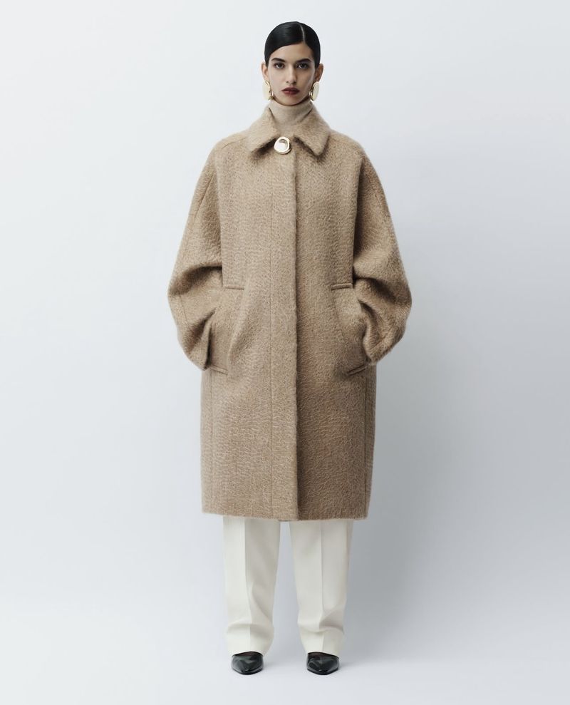 Adult woman wearing a long tan coat, cream turtleneck, and white trousers, standing indoors against a neutral background in a fashion shot.