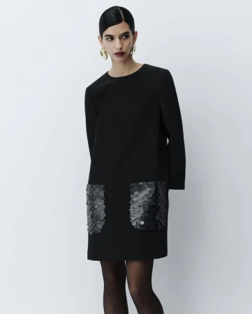 Model wearing a black mini dress with sequined pockets and large gold earrings against a plain light grey background
