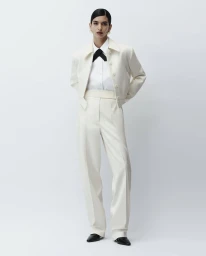Model wearing a cream jacket and trousers with a white shirt and black necktie against a plain white studio backdrop.