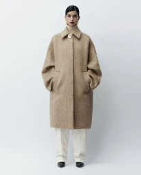 Model wearing a textured camel coat and cream trousers standing against a light grey studio background.