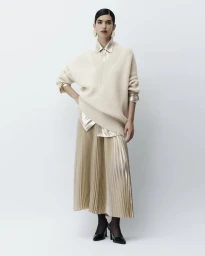 Model Gal Dahan wearing an oversized cream knit sweater layered over a shimmering pleated midi skirt against a pale studio backdrop.