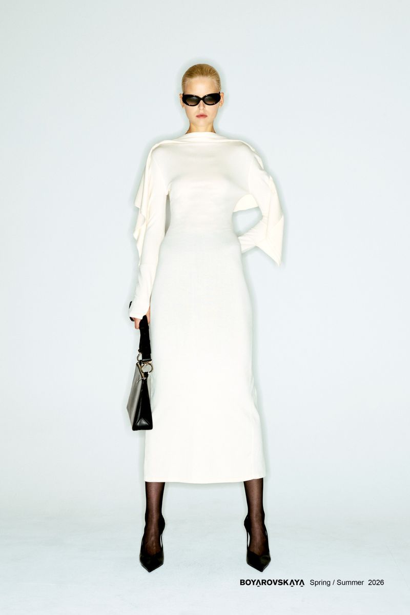 Full-length shot of a woman with fair skin and blonde hair wearing a cream-colored dress, black shoes, and sunglasses against a pale background.