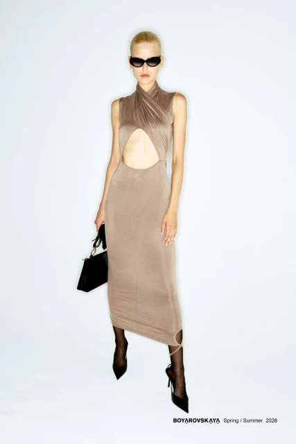 A fashion model poses in a beige dress with black heels and handbag in a studio setting against a bright white backdrop.