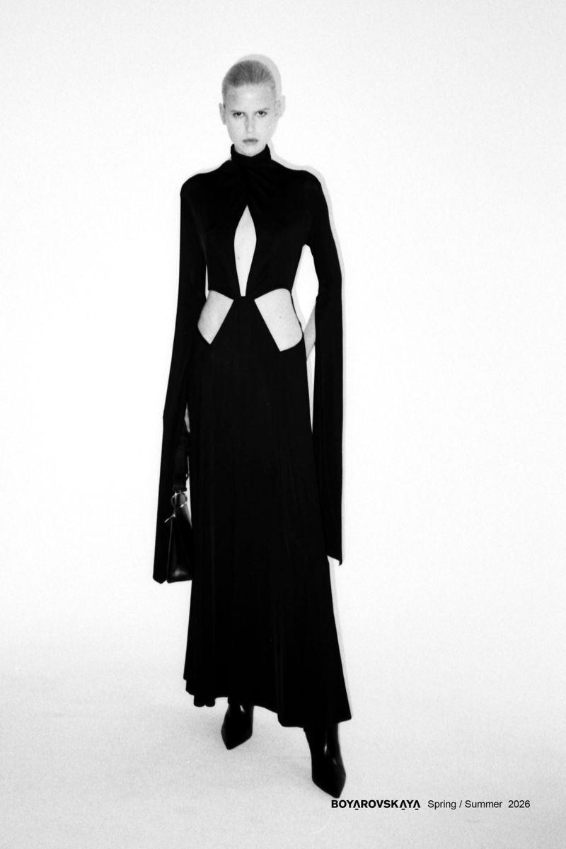 Model in a black cutout long-sleeve dress holding a handbag against a plain white background for Boyarovskaya SS26.