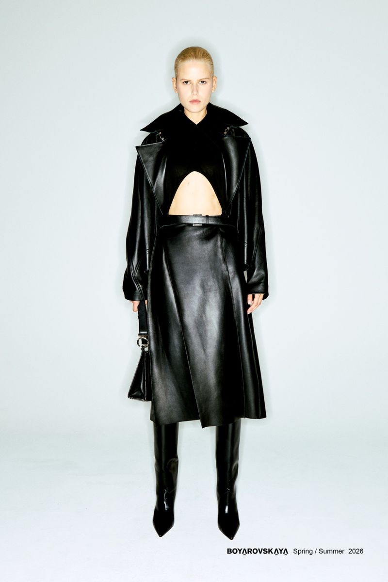 Full-length studio shot of a woman in a black leather coat, skirt, and boots, standing against a white background, fashion editorial.