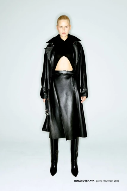 Full-length studio shot of a woman in a black leather coat, skirt, and boots, standing against a white background, fashion editorial.