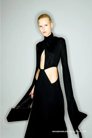 Model in a long black cutout dress with dramatic flared sleeves holding a black handbag against a grey seamless background