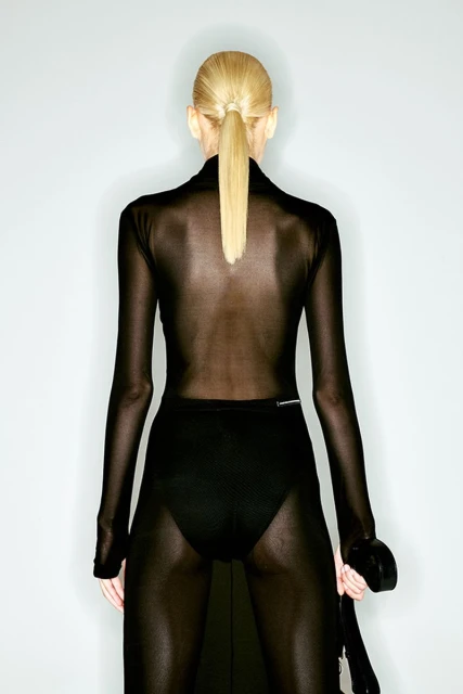 Back view of a slender adult woman with blond hair in a ponytail, wearing a sheer black dress and standing against a pale gray background.