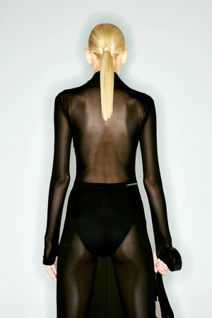 Back view of a slender adult woman with blond hair in a ponytail, wearing a sheer black dress and standing against a pale gray background.