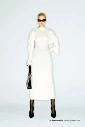 Model in dark sunglasses wearing a long white dress and carrying a black handbag against a light studio backdrop