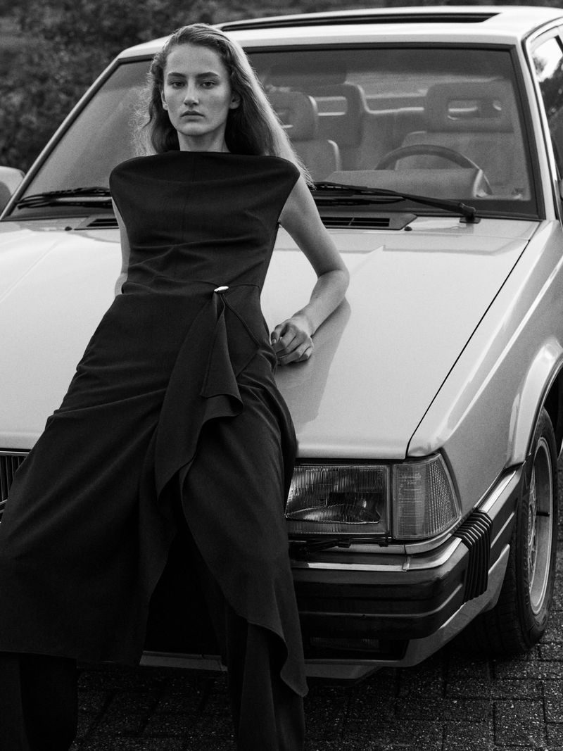Elegant adult female model posing in a black dress, leaning on the hood of a light silver car with a vintage aesthetic in a portrait-style photograph.