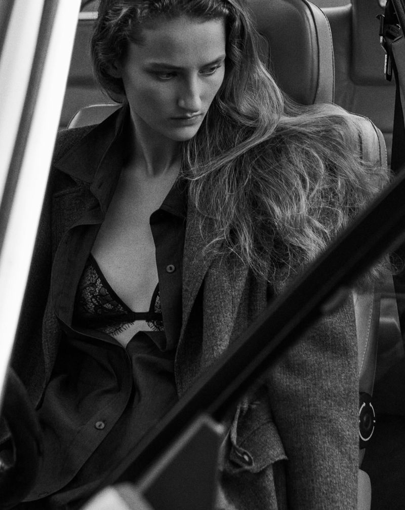 Black and white medium shot of adult female fashion model wearing a blazer and lace bra in the driver seat of a car.