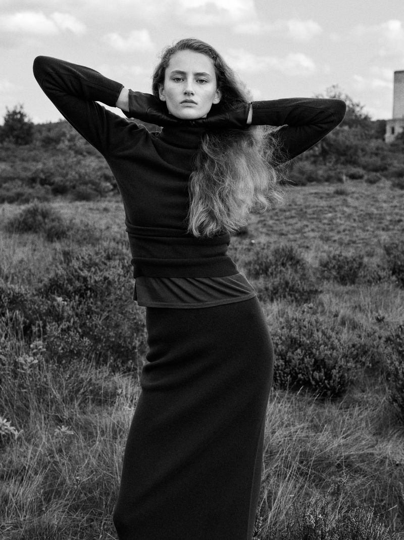 A young woman with long hair is standing in a grassy field wearing a dark turtleneck sweater and skirt, posing with her hands behind her head.