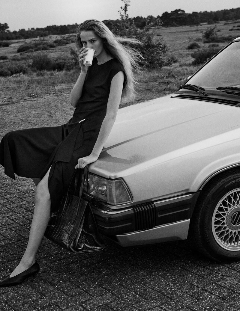 Monochrome fashion shot of a young woman in a dark dress sitting on the hood of a car holding a cup in a rural outdoor setting.