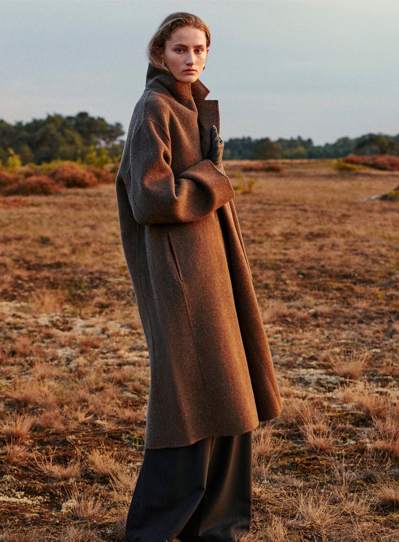 A poised woman stands in a sprawling field wearing a wool coat and dark pants, illuminated by soft light at dusk.