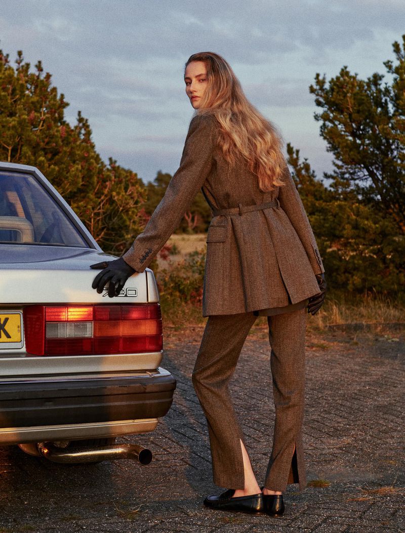 Fashionable adult female model posing in stylish brown tweed suit with classic car and natural outdoor backdrop at dusk or dawn.