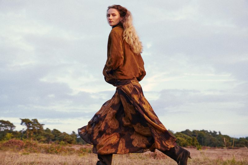 A woman in a brown top and patterned skirt confidently strides through a grassy field with scattered trees under a partly cloudy sky.