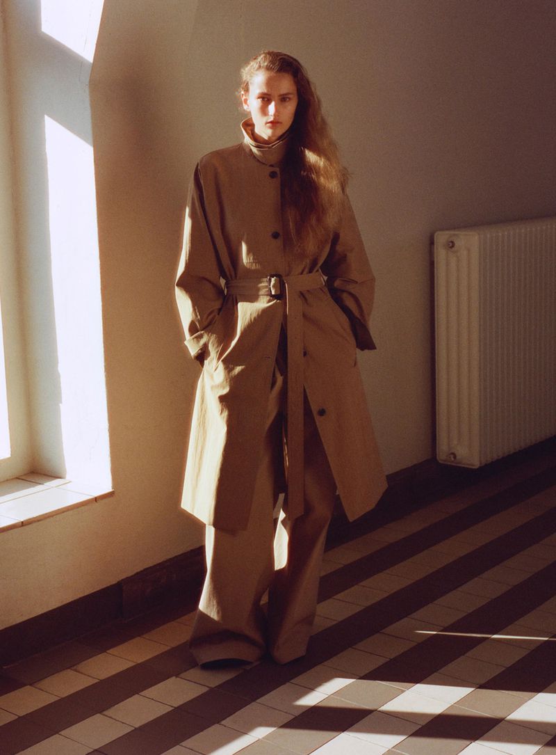 A young woman poses indoors wearing a beige trench coat and matching trousers, bathed in soft sunlight and casting a shadow on the floor.