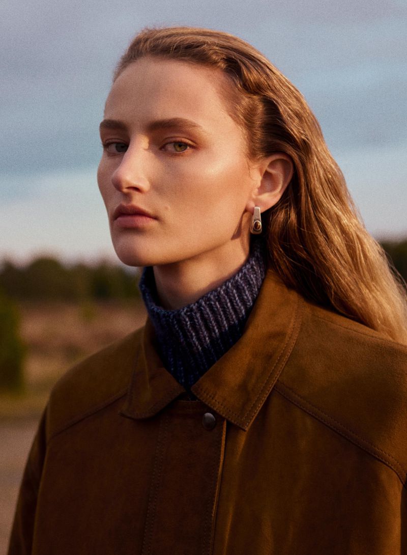 Portrait of a model wearing a brown suede jacket and blue turtleneck against an outdoor landscape background