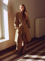 Model in long tan belted coat and trousers standing indoors on striped tile floor with sunlight streaming through a window.