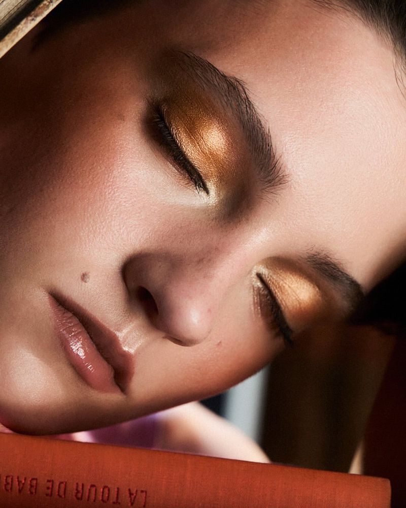 Close-up of a young adult female with closed eyes and gold eyeshadow on a book, beauty and fashion, natural lighting, indoor setting.