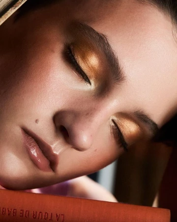 Close-up of a young adult female with closed eyes and gold eyeshadow on a book, beauty and fashion, natural lighting, indoor setting.