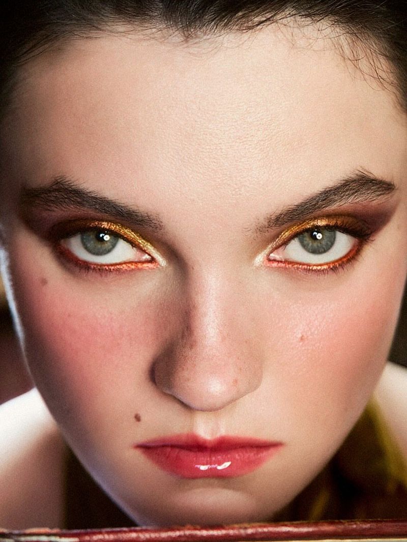 Close-up portrait of a young woman with bold gold and brown eye makeup, full glossy lips, and fair skin.