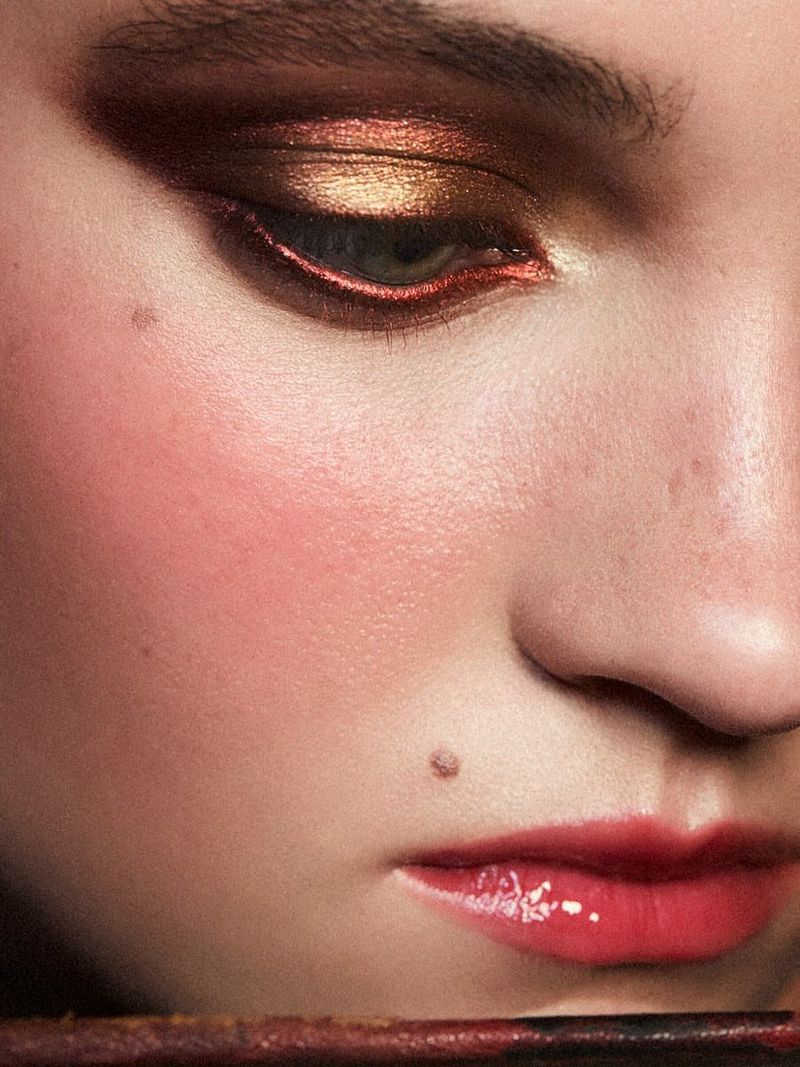 Close-up of a female face showcasing copper and gold eye makeup, rosy cheeks, and glossy red lips. Beauty shot with soft lighting.
