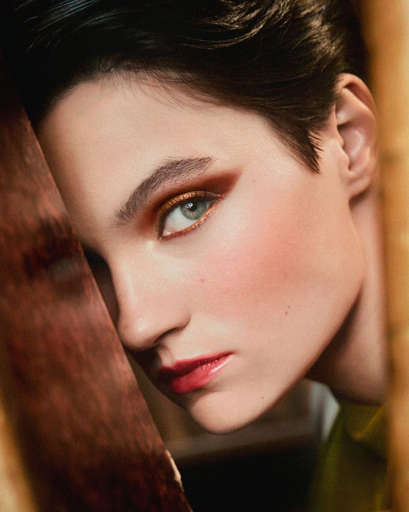 Close-up portrait of a young adult female with short dark hair, brown eyeshadow, and red lipstick, gazing slightly to the left.