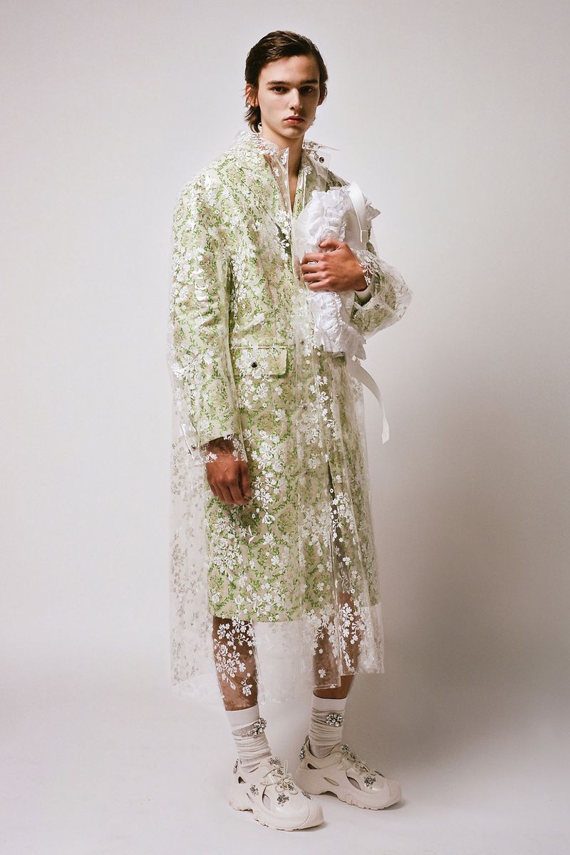 Full shot of a fashion model wearing a green-and-white coat with floral lace over a white shirt and white sneakers in a studio setting.