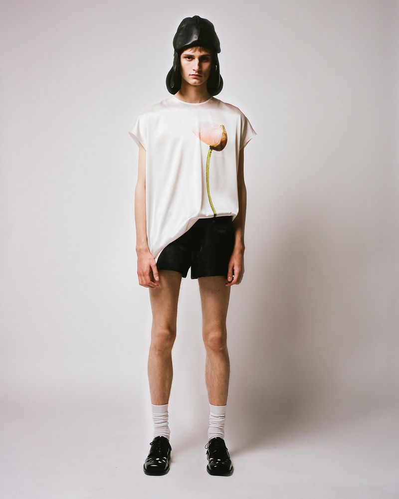 A full-length studio shot of a slender person wearing a cream-colored top, black shorts, white socks, black shoes, and a black hat.