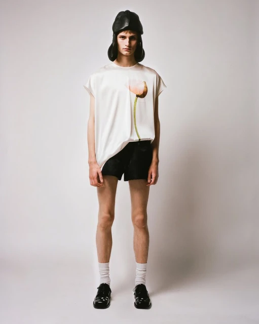 A full-length studio shot of a slender person wearing a cream-colored top, black shorts, white socks, black shoes, and a black hat.