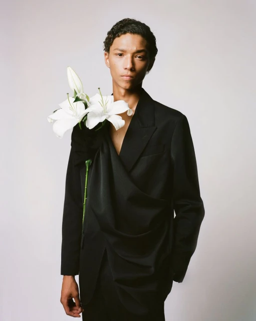 A model poses indoors wearing a draped black suit jacket and pants, accessorized with white lilies for a fashion editorial shoot.